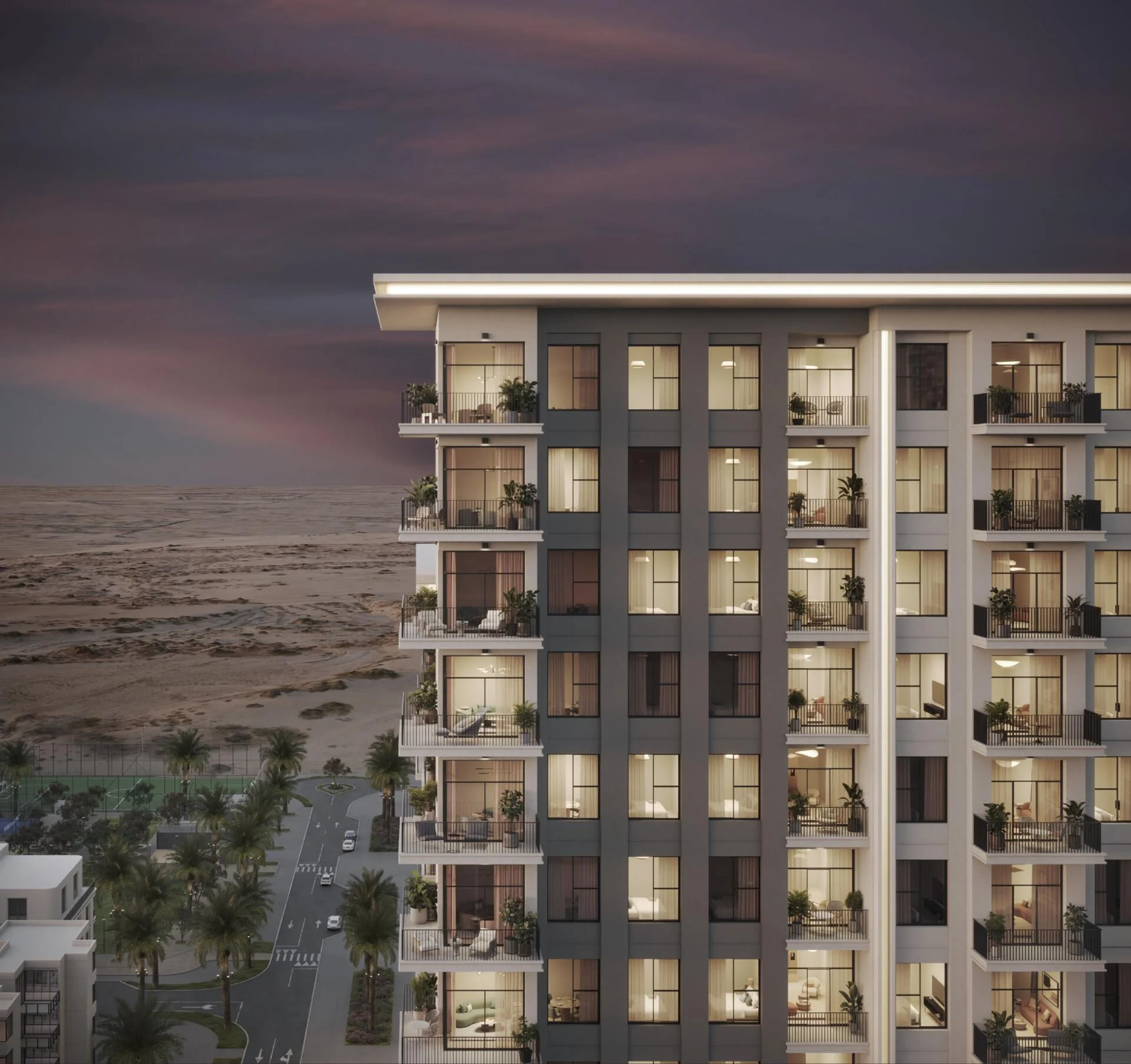 A nighttime view of the modern Elaya apartment building with lit windows, overlooking a road lined with palm trees and a sandy landscape under a cloudy sky.